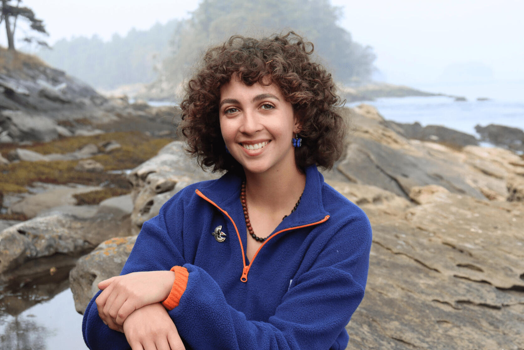 Olivia deBourcier sitting on rocks by the ocean wearing a blue sweatshirt.